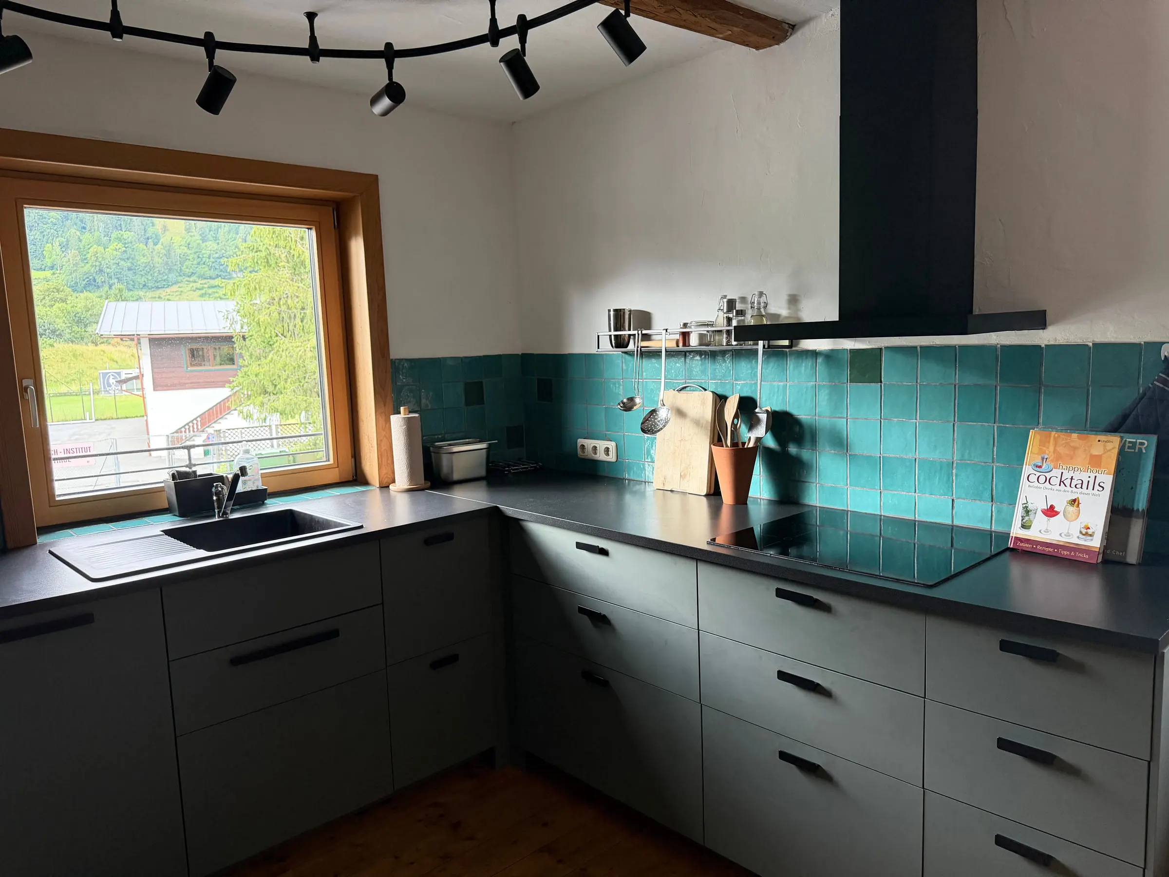 Küche über Eck mit salbeigrünen Fronten, dunkler Arbeitsplatte, grünen Fliesen und großem Fenster