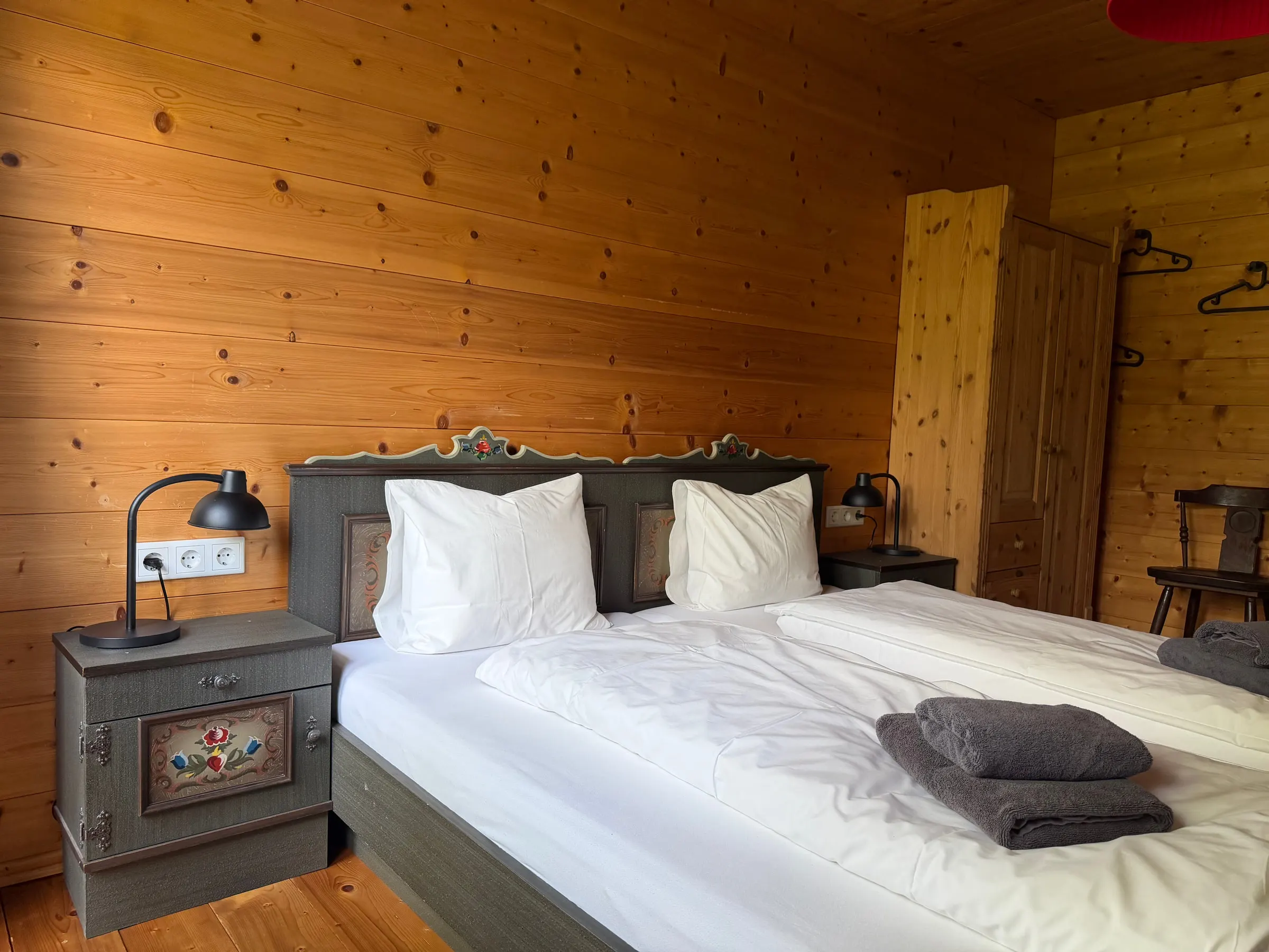 Schlafzimmer mit bemaltem grauen Bauernbett, zwei Nachtkästchen mit Leselampen und heller Holzwand