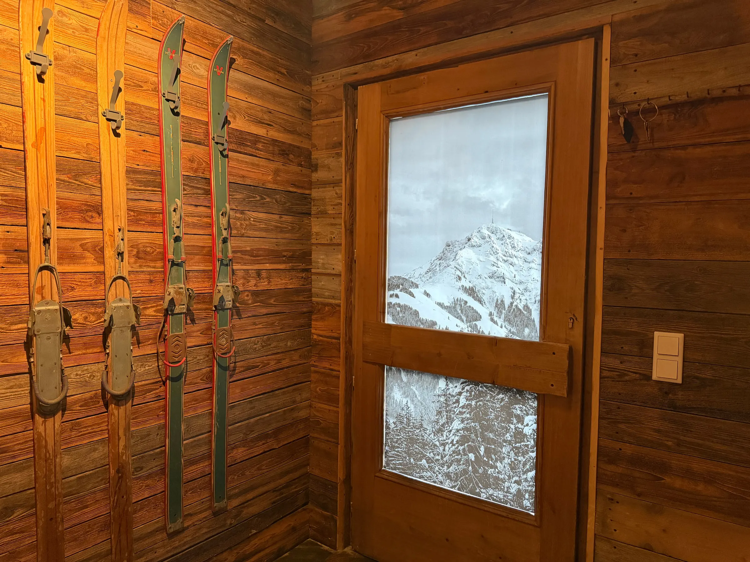Vorraum mit alten Holzski an der Wand und einer Glastür mit Blick auf verschneite Berge