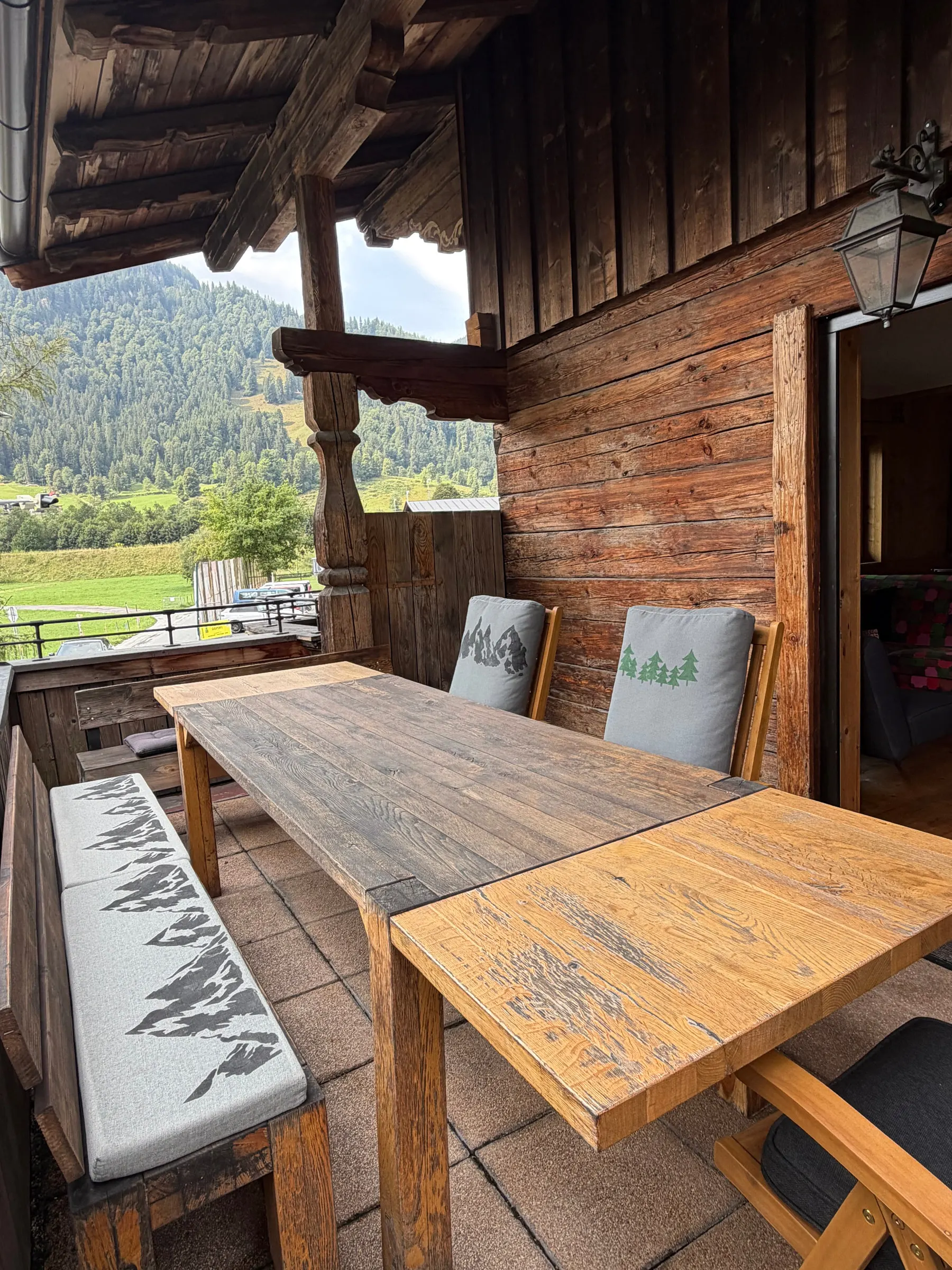 Überdachte Terrasse mit langem Holztisch, Bank und Stühlen, dahinter bewaldete Berghänge