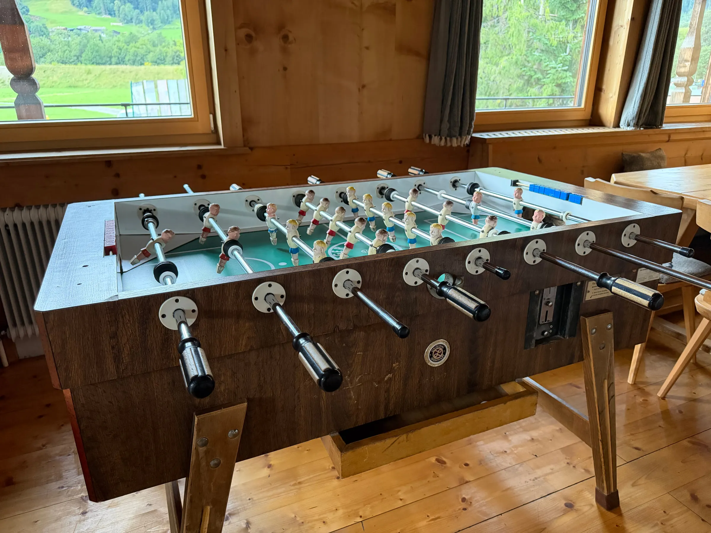 Tischkicker aus dunklem Holz vor zwei Fenstern mit Blick auf Wiesen und Berge