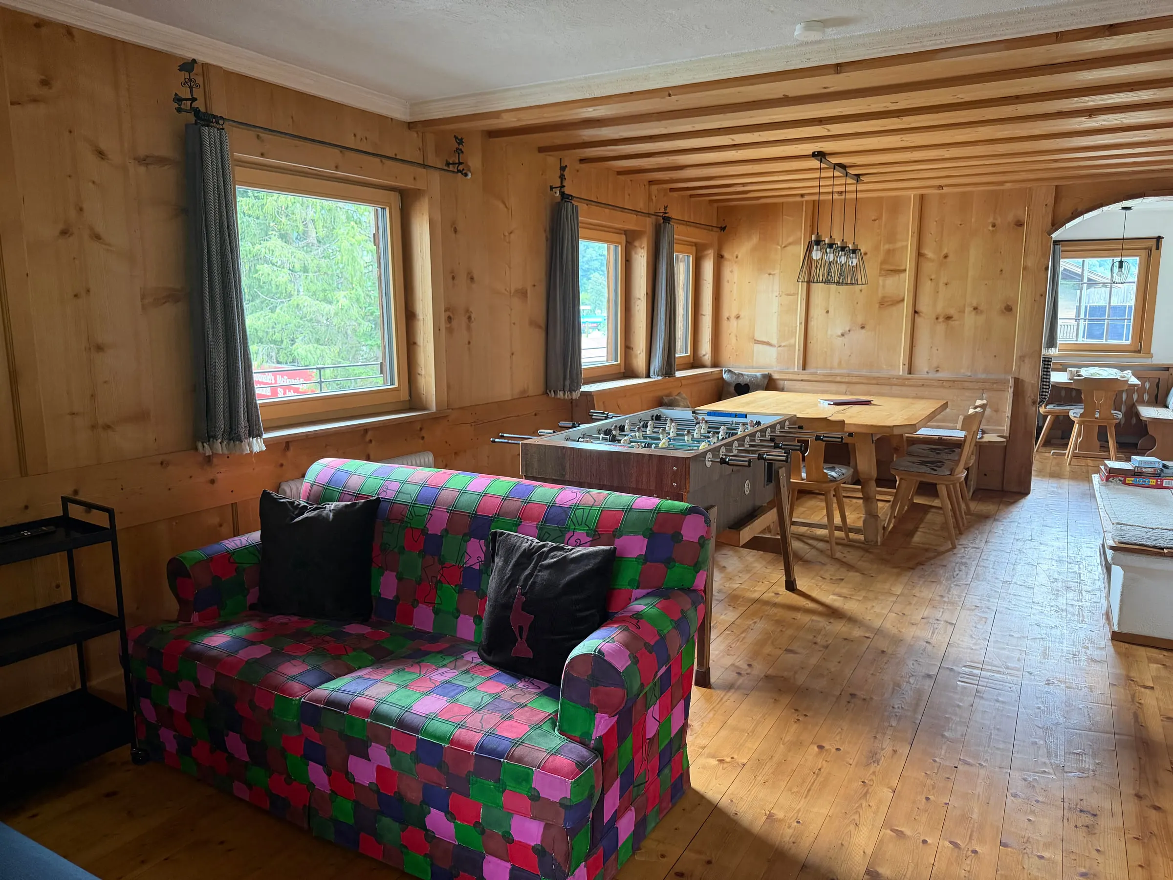 Wohn-Esszimmer mit Tischkicker, langem Holztisch, buntem Sofa und drei Fenstern zum Tal